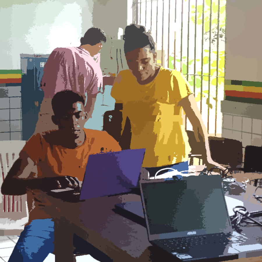 Na imagem podemos ver dos jovens. O rapaz está sentado enuanto utiliza um laptop que está sobre a mesa. Ao seu lado, se encontra uma jovem de pé, observado sua ação. Sobre a mesa há dois laptops.